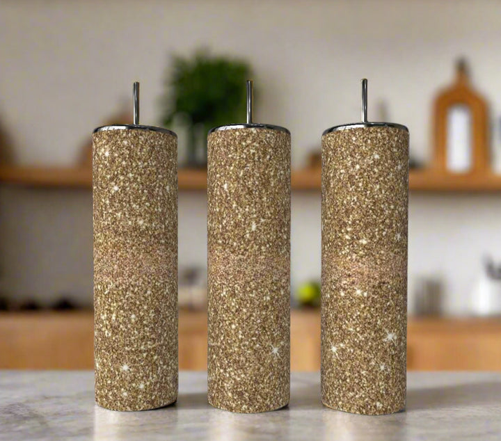 Three cylindrical spice shakers with brown, sand-like texture and silver tops are lined up on a kitchen counter next to the Gold Fearless Shimmer 20oz Skinny Tumbler by Kreative Kreationz, adding a touch of luxury drinkware to the scene.