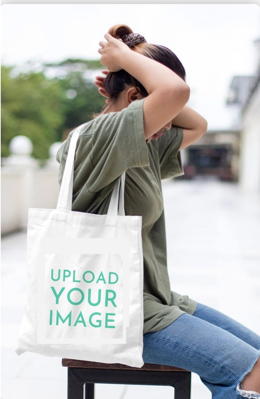Outdoors, a person ties their hair back while wearing a green t-shirt, ripped jeans, and carrying the Tote-ally Yours Canvas Bag by Kreative Kreationz—an eco-friendly, roomy tote printed with "UPLOAD YOUR IMAGE.