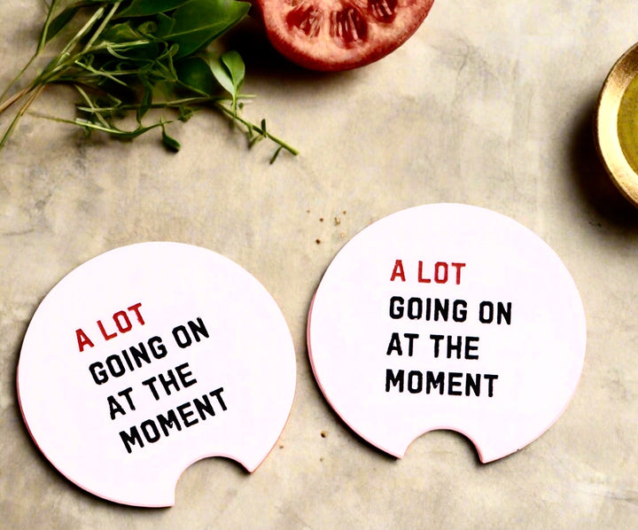 Two RED Car Coasters by Kreative Kreationz, each 2.7" round ceramic, rest on a marble surface with "A LOT GOING ON AT THE MOMENT" text, beside herbs, half a tomato, and a small bowl.