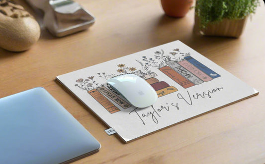 A Taylor’s Version Mousepad by Kreative Kreationz with a floral and book design, including titles like "Fearless," "Speak Now," and "Red" (all marked as "Taylor's Version"), enhances a wooden desk. Ideal for Swiftie gifts, it's paired with a wireless mouse and part of a laptop.
