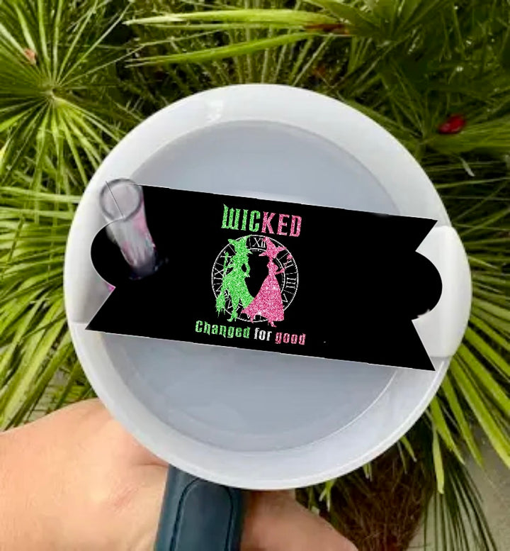 A hand holds a white cup with the Kreative Kreationz Wicked - Stanley Lid Topper and a black straw tag saying "WICKED Changed for good" in green and pink with character silhouettes; green palm leaves appear in the background.