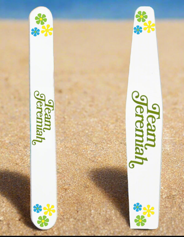 Two Kreative Kreationz Team Jeremiah Ceramic Nail Files featuring green text and colorful flowers stand upright on sandy beach, with a blurred blue sky or water in the background.