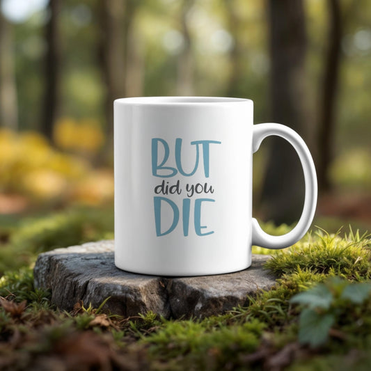 The But Did You Die Point Mug by Kreative Kreationz features "BUT did you DIE" in vibrant colors on a white, dishwasher-safe mug, pictured on a forest tree stump surrounded by moss and sunlight.