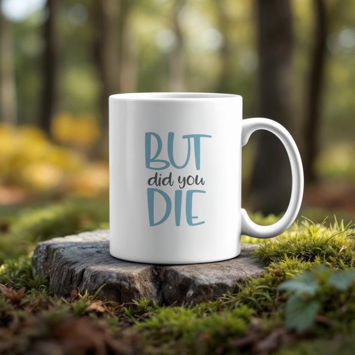 The But Did You Die Point Mug by Kreative Kreationz features "BUT did you DIE" in vibrant colors on a white, dishwasher-safe mug, pictured on a forest tree stump surrounded by moss and sunlight.
