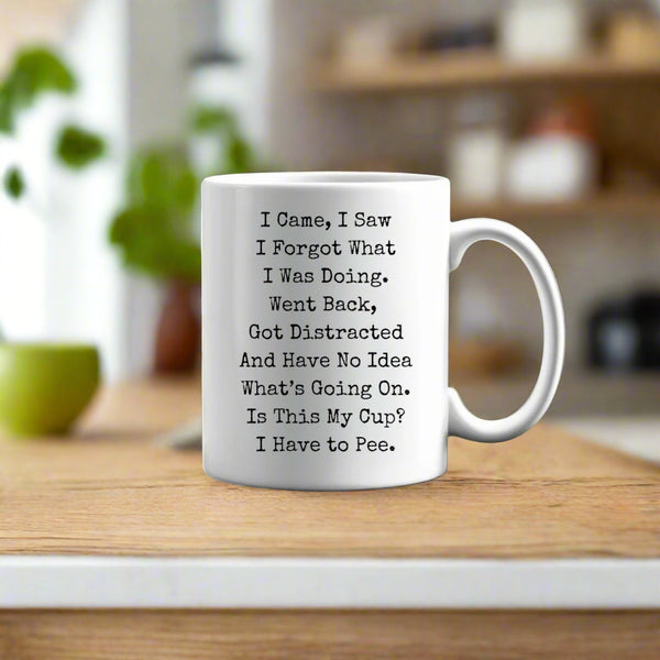 The ADHD Ceramic Mug by Kreative Kreationz, a white dishwasher-safe mug with funny black text—"I came, I saw I forgot what I was doing. Went back, got distracted and have no idea what's going on."—rests on a tree stump in a sunlit forest.