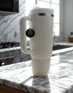A large white insulated mug with a handle rests on a marble countertop in sunlight. Hanging from its lid is the Kreative Kreationz REP|13 Snake Engraved Cup Charm featuring a coiled snake and 