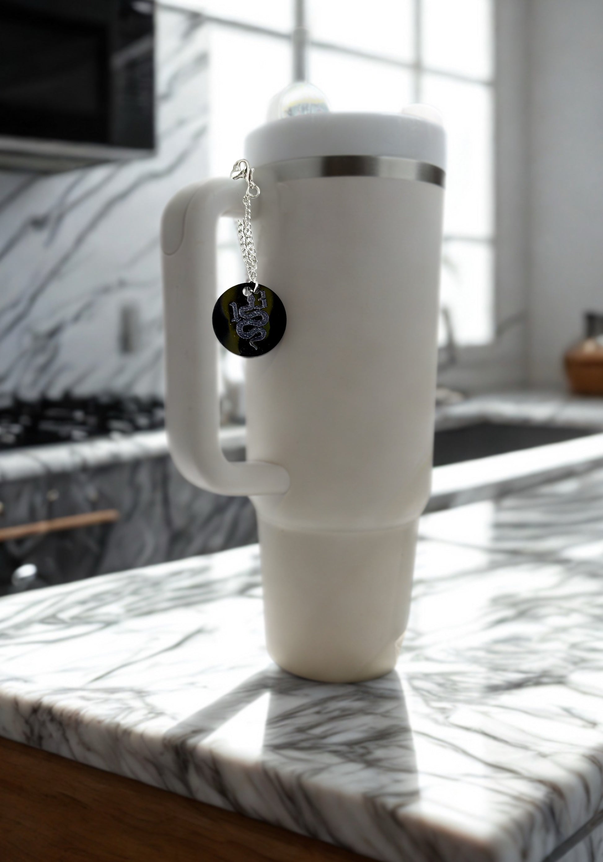 A large white insulated mug with a handle rests on a marble countertop in sunlight. Hanging from its lid is the Kreative Kreationz REP|13 Snake Engraved Cup Charm featuring a coiled snake and "Don't Tread On Me.