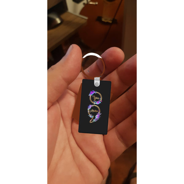 A hand holds a black rectangular "You Matter Keychain" by Kreative Kreationz, featuring a circular floral design and subtle semicolon to promote positivity and mental health awareness.