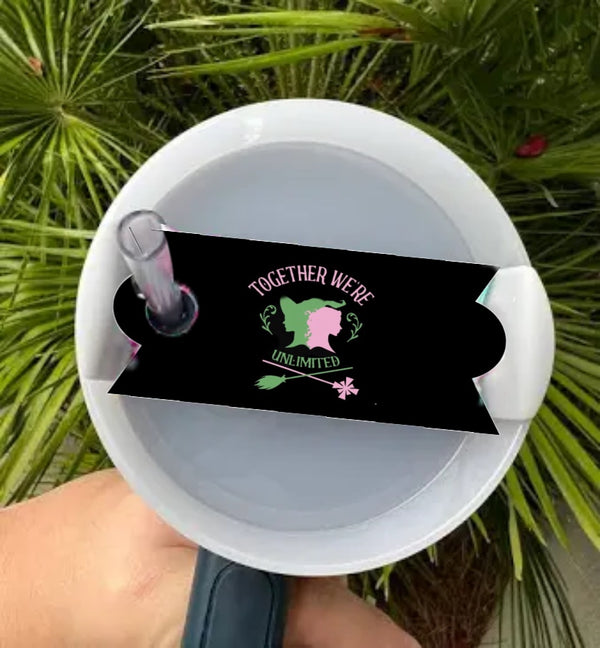 A hand holds a white tumbler with a straw and the Wicked -Stanley Lid Topper by Kreative Kreationz. A black sticker features a broom, wand, “TOGETHER WE’RE UNLIMITED,” and two face silhouettes. Green palm leaves are in the background.