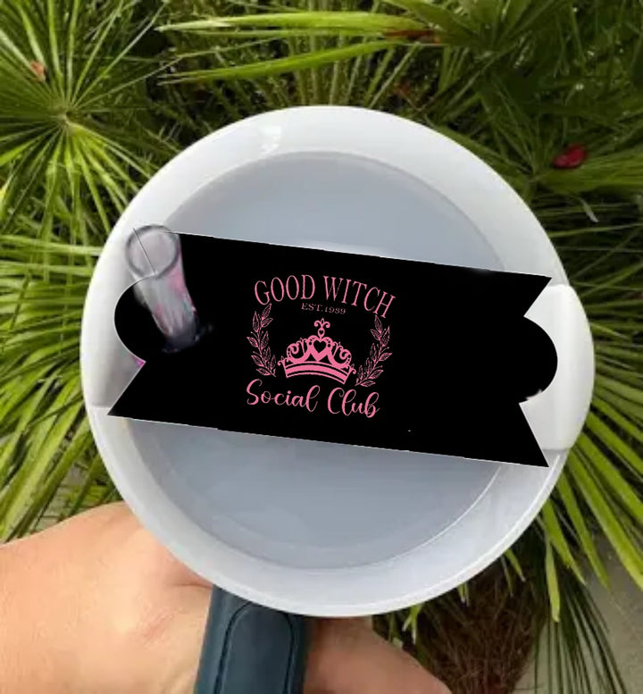 A hand holds a durable tumbler accessory from Kreative Kreationz: the Wicked | Good Witch Social Club Stanley Lid Topper, designed like a white mug with a black ribbon, pink "Good Witch Social Club" text, crown, and floral accents. Green palm leaves create an enchanting backdrop.